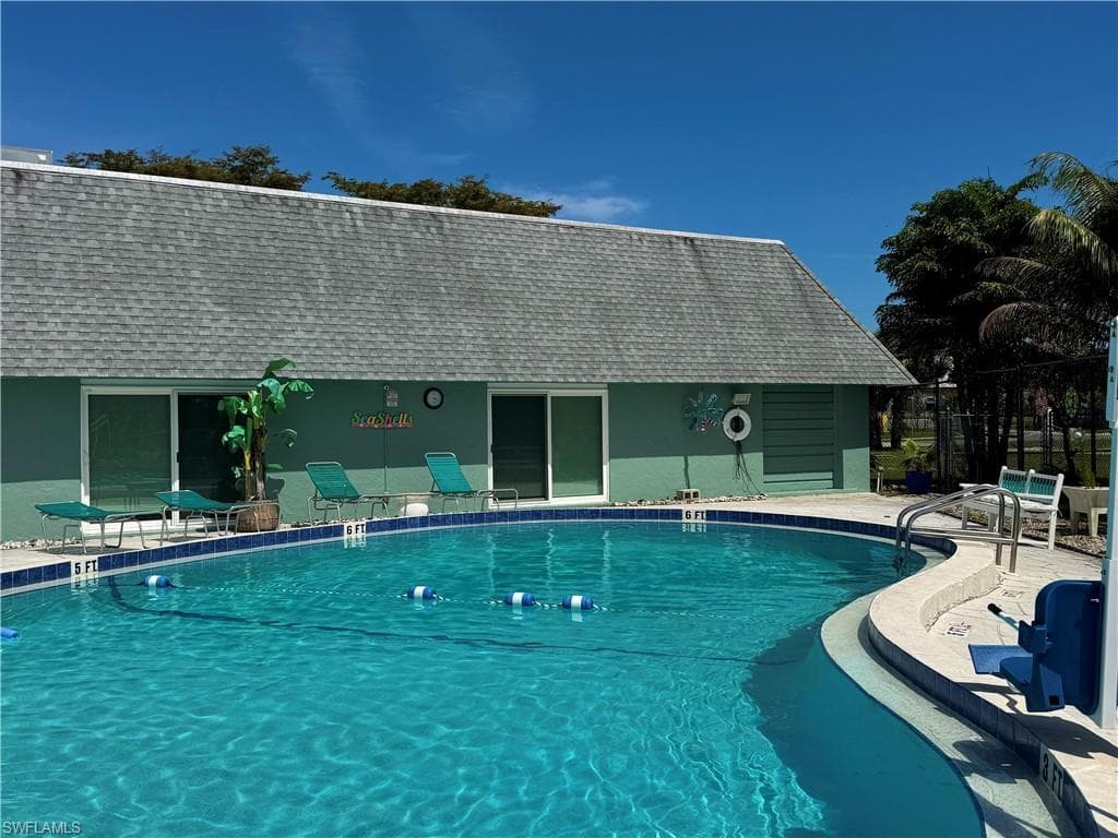 Curved swimming pool with a tile border and light-colored coping, complemented by a green-sided structure featuring multiple sliding glass doors and a gray shingled roof