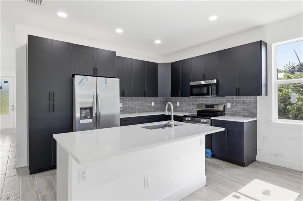 Modern kitchen featuring dark cabinetry, stainless steel appliances, a white island with an integrated sink, a gray subway tile backsplash, and light wood-finish flooring
