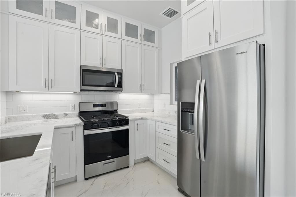 Kitchen with stainless steel appliances, light marble finish floors, white cabinets, light stone counters, and glass insert cabinets VIRTUALLY EDITED