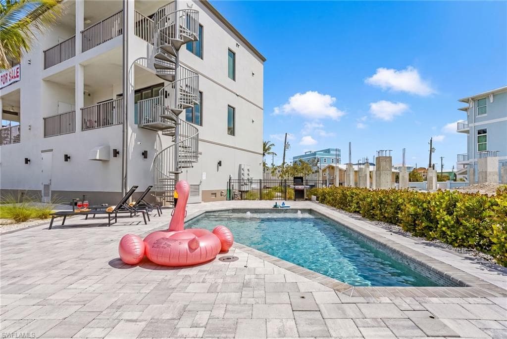 Outdoor living area featuring a rectangular swimming pool with tiled coping, a surrounding paver patio, and a three-story exterior spiral staircase - Virtually Edited Image
