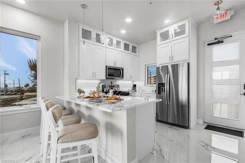 Modern kitchen featuring white cabinetry, a central island with a stone countertop, stainless steel appliances, and large format marble-look tile flooring - Virtually Edited Image