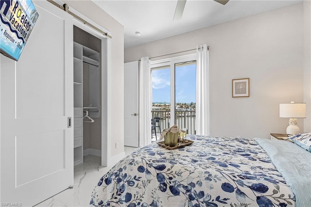 Bedroom featuring white barn door entry to a walk-in closet with built-in shelving, glass sliding doors opening to an outdoor balcony, white-finish flooring, and a ceiling fan - Virtually Edited Image
