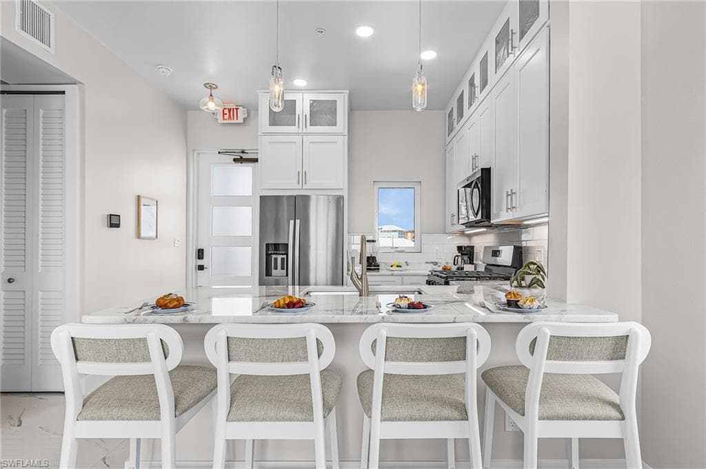 Open-concept kitchen featuring a white subway tile backsplash, stainless steel appliances, white shaker cabinetry with glass-front uppers, and a kitchen island with a light-toned countertop - Virtually Edited Image
