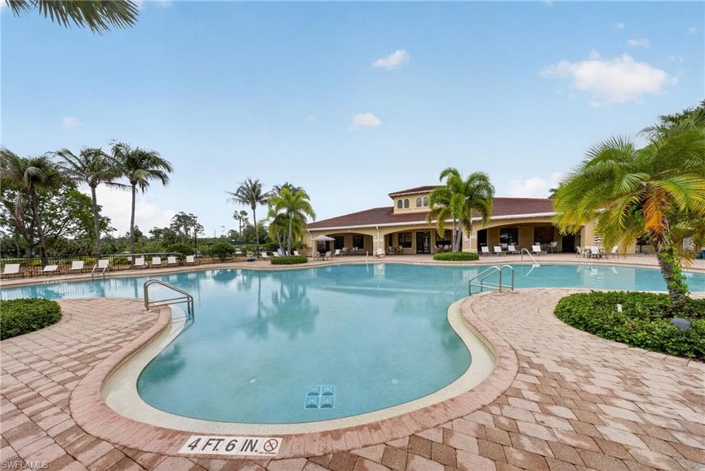 Expansive outdoor swimming pool with a brick paver deck, surrounded by tropical landscaping and a clubhouse building