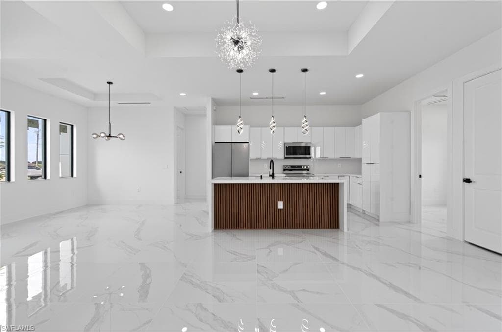 Open-concept living space with large format white tile flooring, a contemporary kitchen island featuring wood paneling and pendant lighting, white high-gloss cabinetry, and stainless steel appliances