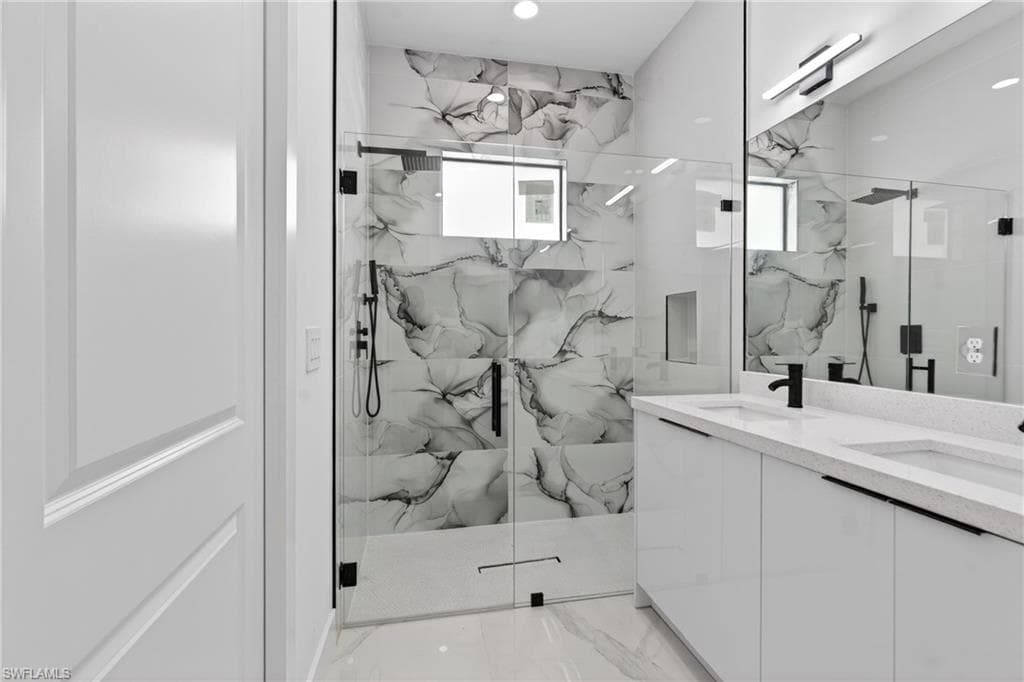 Bathroom featuring a frameless glass shower enclosure with black hardware, large format stone-look tile, a dual vanity with white cabinetry, a light-toned countertop, and matte black plumbing fixtures