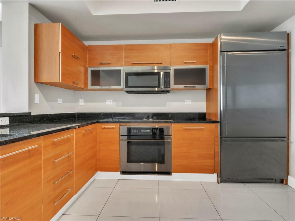 Kitchen featuring appliances with stainless steel finishes, light tile patterned flooring, dark stone countertops, and brown cabinets