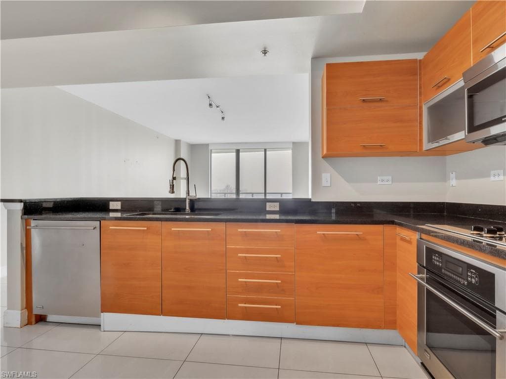 Kitchen featuring brown cabinetry, stainless steel appliances, light tile patterned floors, dark stone counters, and modern cabinets
