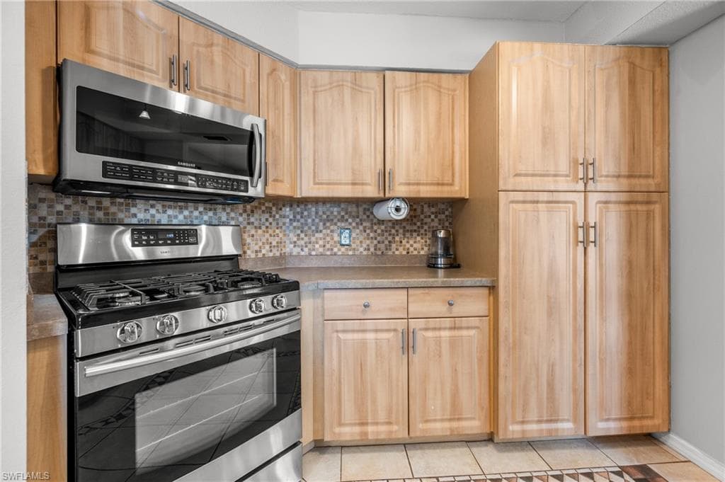 Kitchen featuring wood-finish cabinetry with silver-tone hardware, stainless steel gas range with overhead microwave, a mosaic tile backsplash, and light-toned countertops