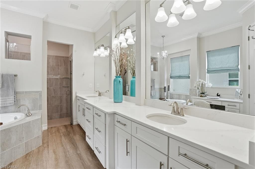 Dual vanity with white cabinetry and integrated sinks set beneath large mirrors, wood-finish flooring, a tiled corner soaking tub, and a shower with glass enclosure