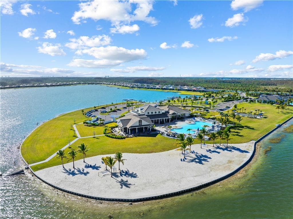 Expansive community amenity island featuring a large clubhouse, resort-style swimming pool, sandy beach area with palm trees, and manicured lawns