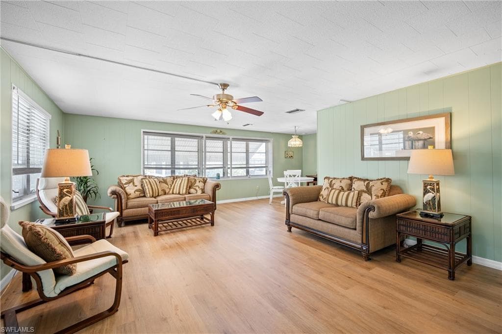 Spacious living area featuring wood-finish flooring, light green walls, multiple windows with blinds, a ceiling fan with integrated lighting, and a defined dining area beyond