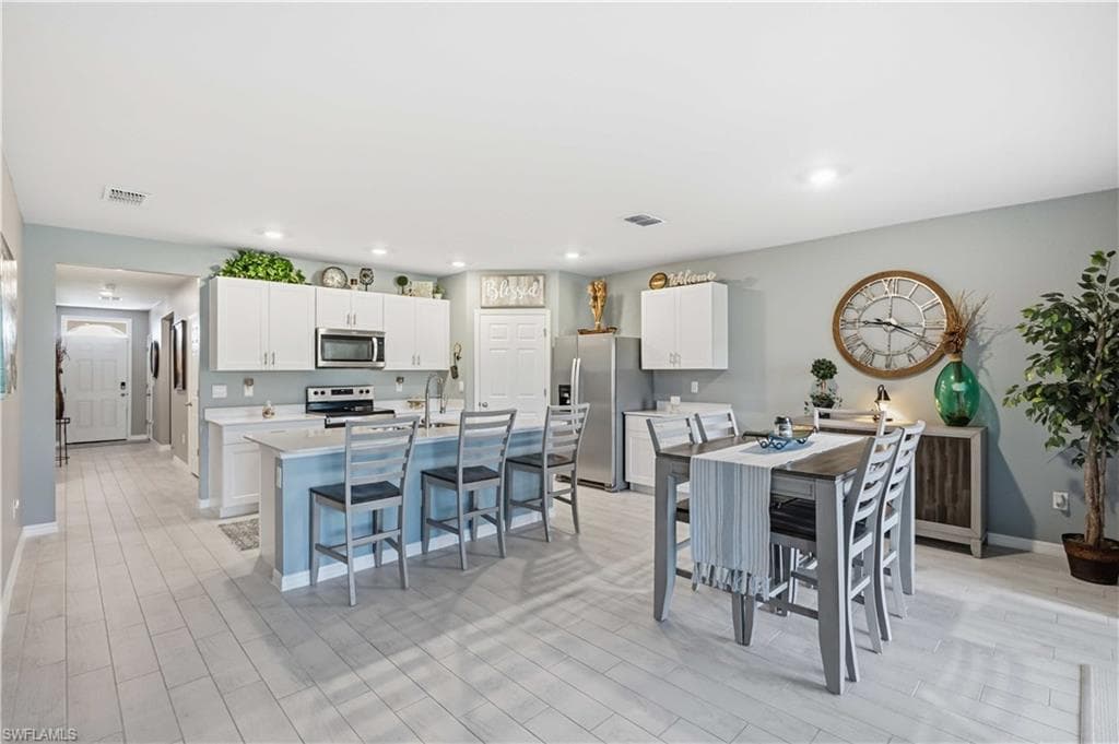 Open-concept kitchen and dining area featuring light-toned flooring, white upper cabinetry, a central island with bar seating, stainless steel appliances, and recessed lighting