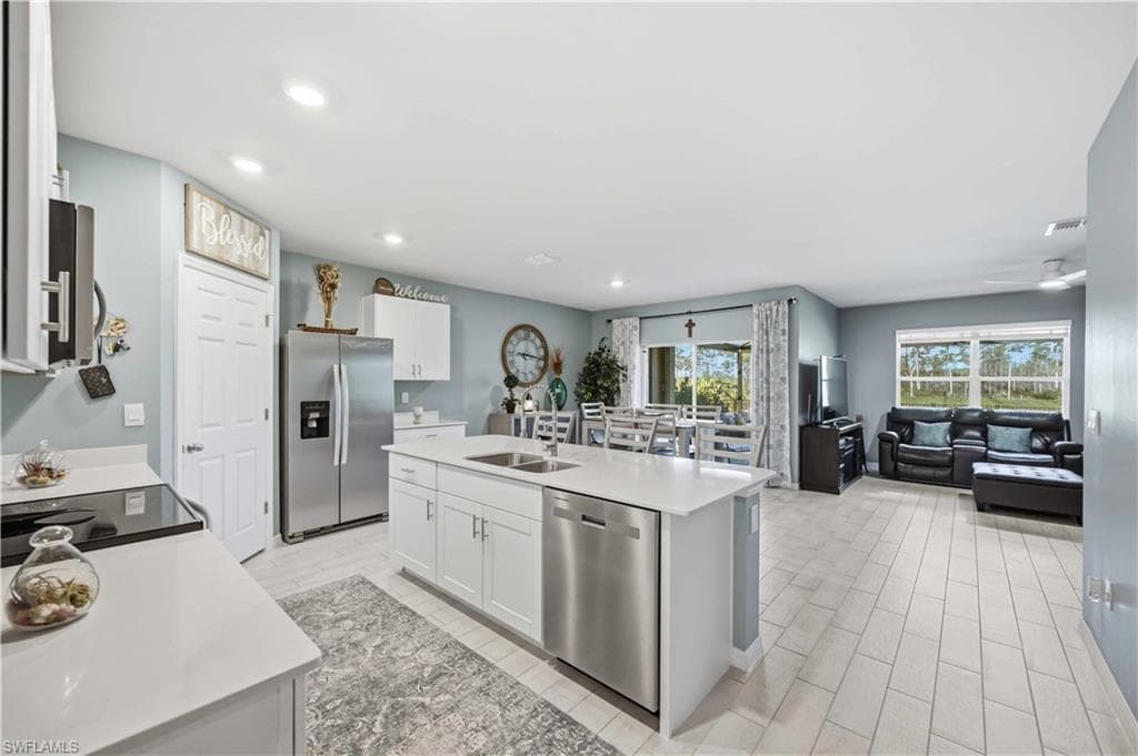Open-concept living space featuring a central kitchen island with a double basin sink, white cabinetry, integrated stainless steel appliances, and light-toned tile flooring