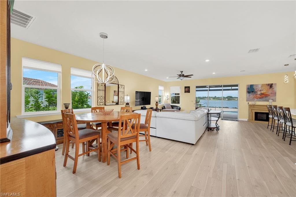 Expansive open-concept living area featuring light wood-finish flooring, a contemporary ceiling fan, recessed lighting, a built-in fireplace, and sliding glass doors opening to waterfront scenery