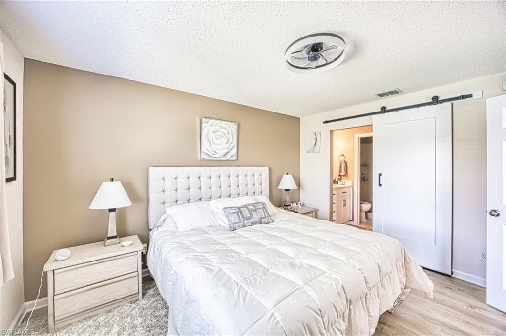 Bedroom with a barn door, a textured ceiling, light wood finished floors, ensuite bathroom, and baseboards