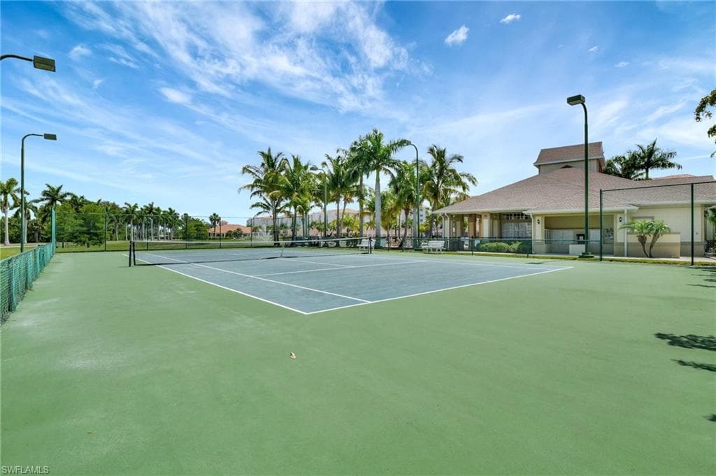 Full-size tennis court with professional netting, surrounded by mature palm trees and green landscaping, featuring perimeter fencing and court lighting