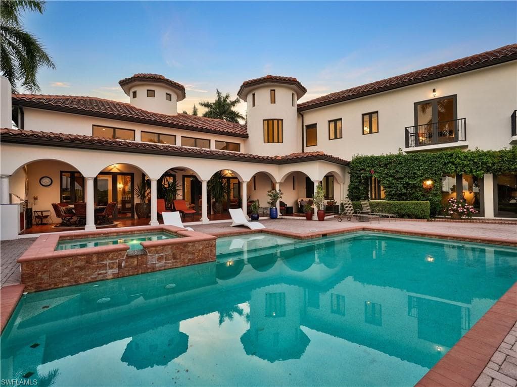 Mediterranean-style exterior featuring a spacious pool, integrated spa with stone detailing, extensive tiled patio, arched covered patio, and a second-story balcony with iron railing