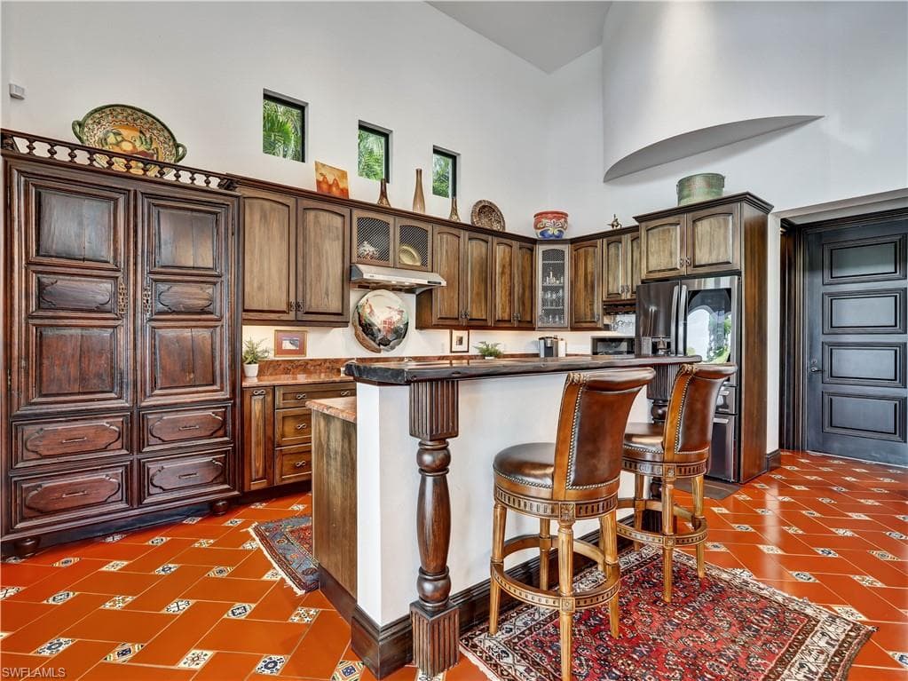 Kitchen featuring terracotta tile flooring with decorative accents, custom wood cabinetry, a built-in refrigerator, a breakfast bar with a decorative newel post, and high ceilings