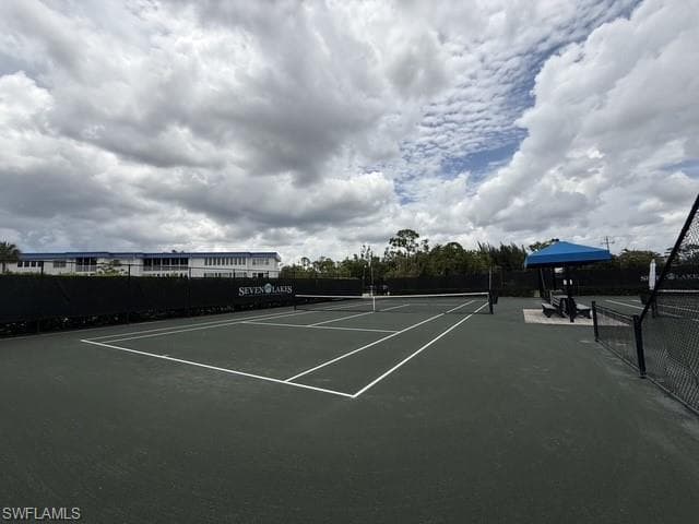 Tennis court featuring a dark green playing surface with white lines, black perimeter fencing, a blue shaded seating area, and lush green landscaping