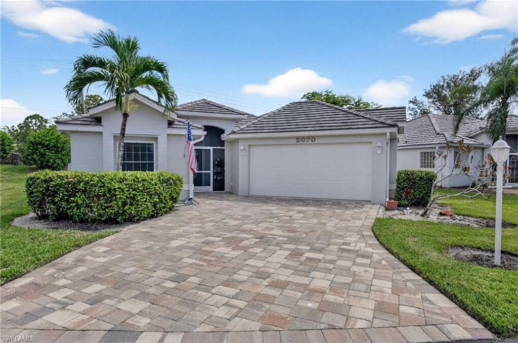 Exterior residence featuring a paver driveway, an attached two-car garage, a tiled roof, mature landscaping, and a screened entry