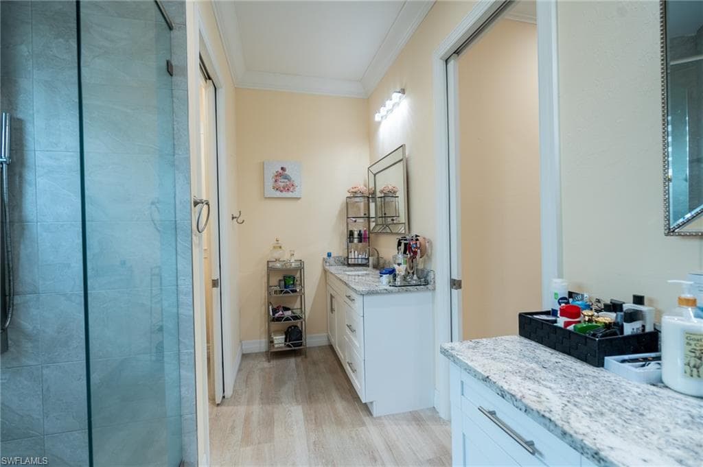 Bathroom featuring a glass-enclosed shower with large format tile, wood-finish flooring, dual vanities with light-toned countertops, and crown molding