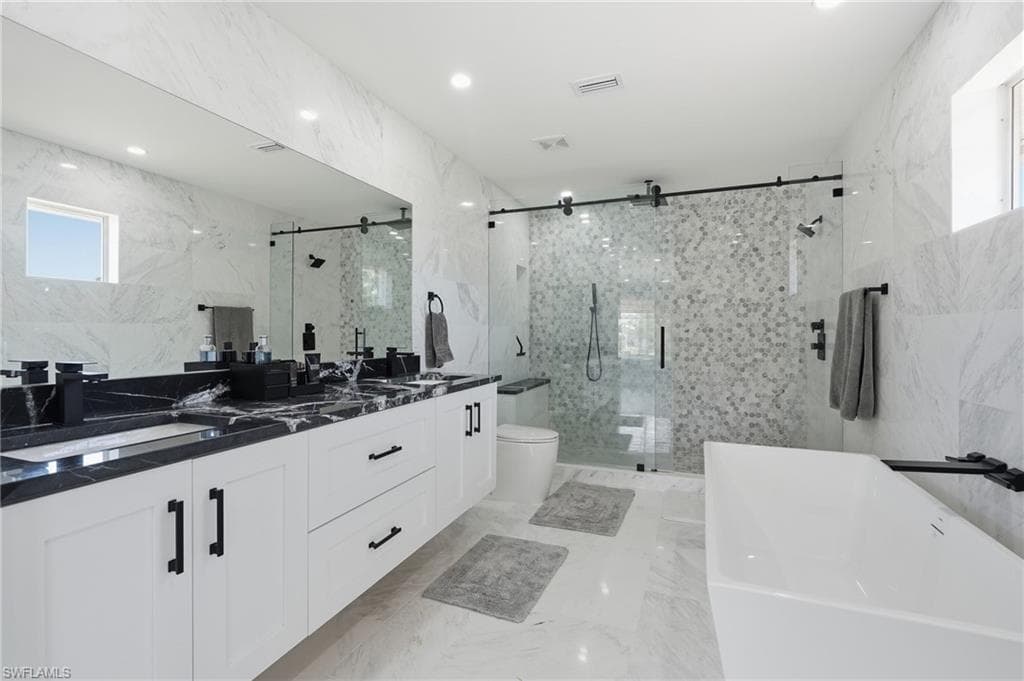 Spacious bathroom featuring a dual vanity with black countertops, a freestanding soaking tub, a walk-in shower with mosaic tile, and marble-look tile flooring and walls