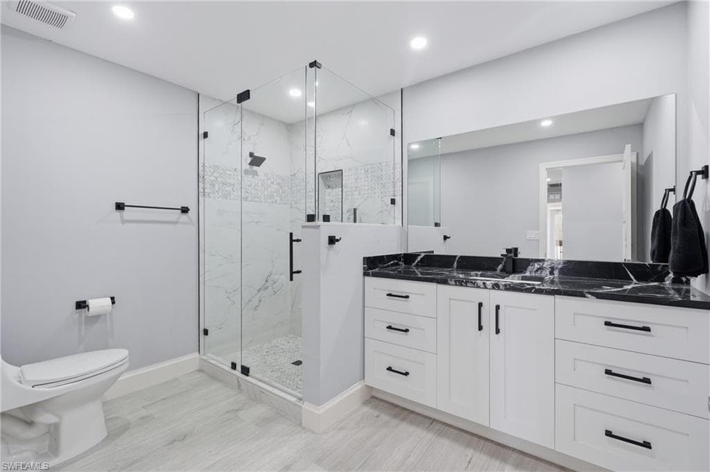 Contemporary bathroom featuring a spacious glass-enclosed shower with white marble-look tile, a dual vanity with a black stone countertop, white shaker-style cabinetry, and light wood-finish flooring