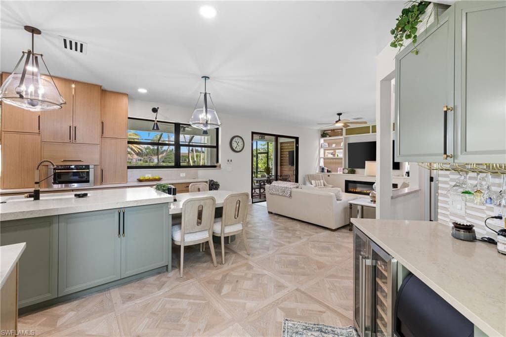 Open-concept interior featuring light wood-finish flooring throughout, a kitchen island with light green cabinetry, integrated appliances, and a built-in wine refrigerator