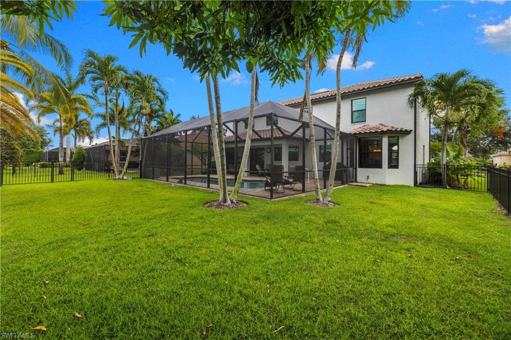 Expansive backyard featuring a screened-in pool enclosure, mature palm trees, a tiled roofline, and a black wrought iron fence