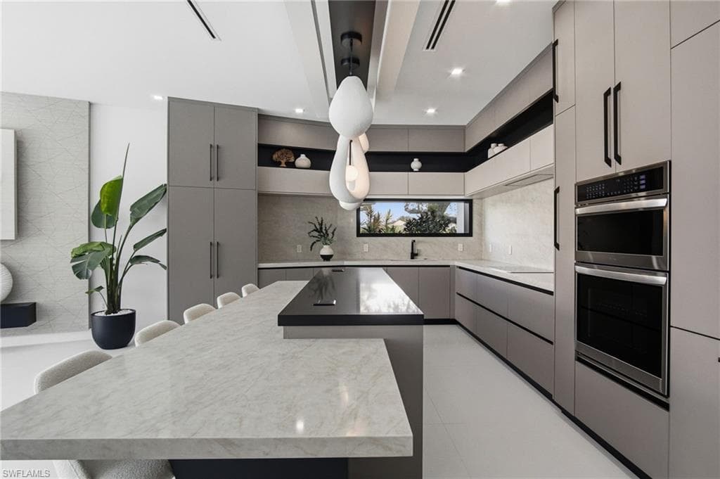 Contemporary kitchen featuring an oversized island with a light-toned countertop, sleek flat-panel cabinetry, integrated double wall ovens, a linear window, and recessed lighting