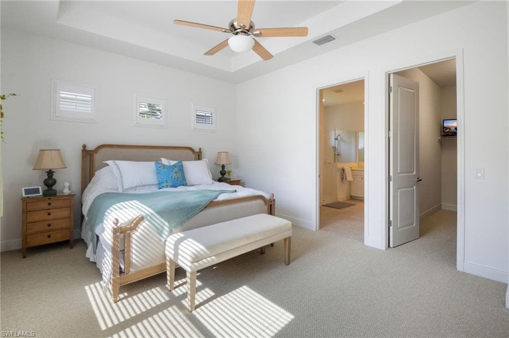 Carpeted bedroom featuring a tray ceiling with a ceiling fan, three windows with white plantation shutters, an open doorway revealing a bathroom with a vanity and shower, and a second open doorway