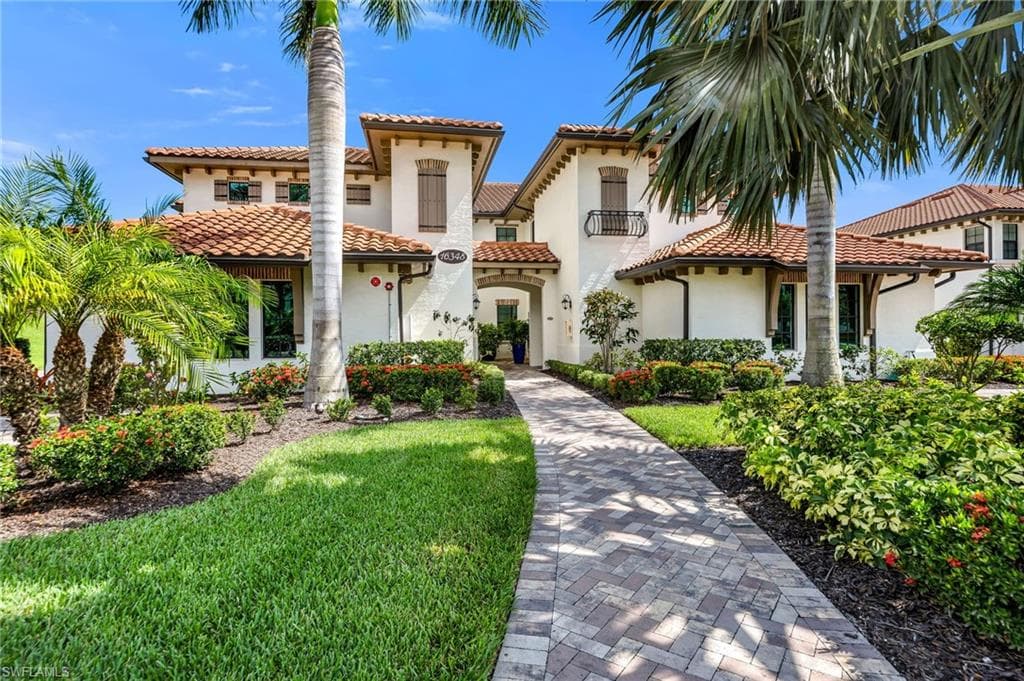 Mediterranean-inspired exterior featuring a paver walkway, mature palm trees, lush landscaping, and a terra cotta tile roof