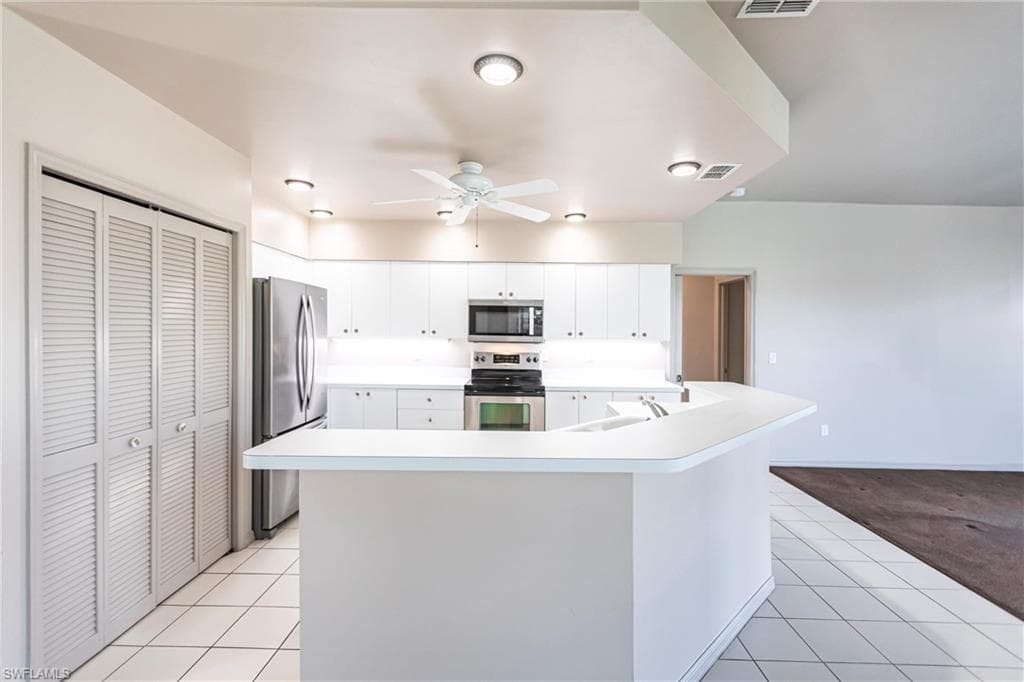Kitchen with white cabinetry, spacious pantry, appliances with stainless steel finishes, a kitchen island with sink, and light tile patterned floors leading to carpeted entertainment area overlooking lake.