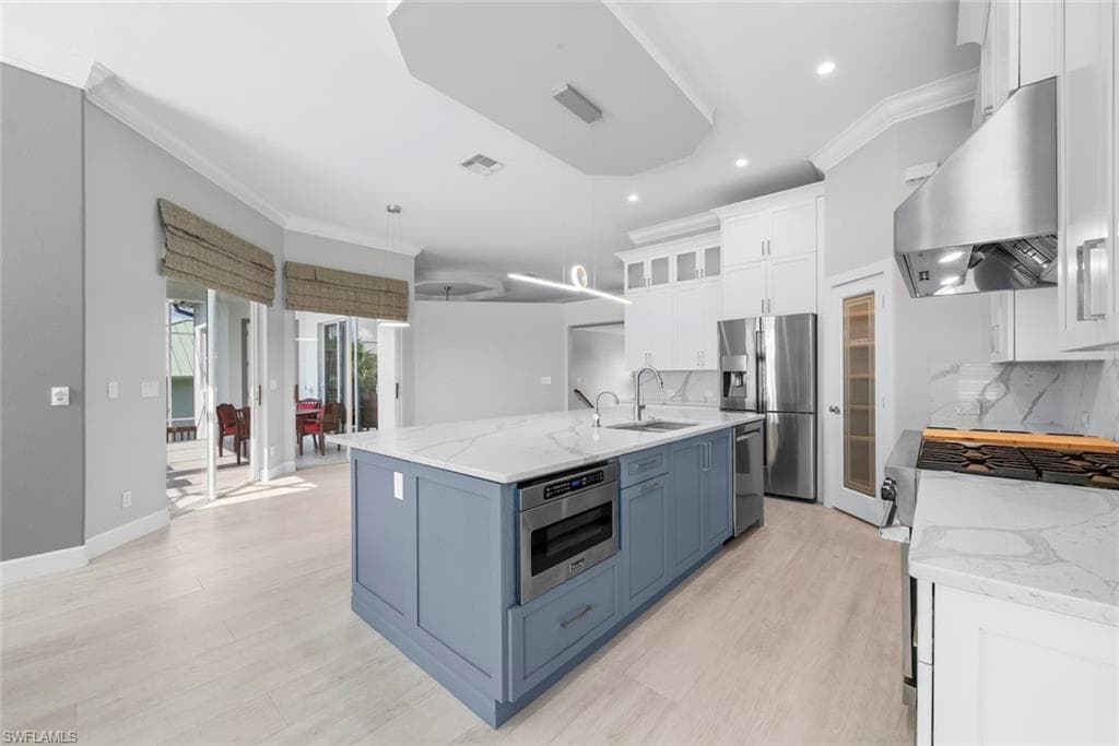 Spacious kitchen featuring an expansive central island with a built-in microwave, light wood-finish flooring, white upper cabinetry, stainless steel appliances, and quartz countertops with a matching backsplash