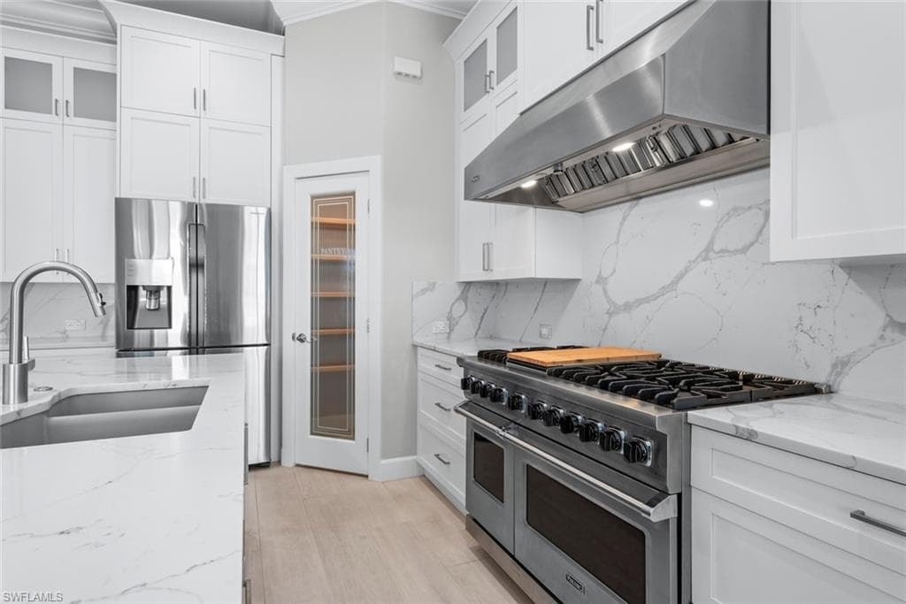 Gourmet kitchen featuring a professional-grade gas range with stainless steel hood, extensive white cabinetry, light wood-finish flooring, and a glass-paneled pantry door