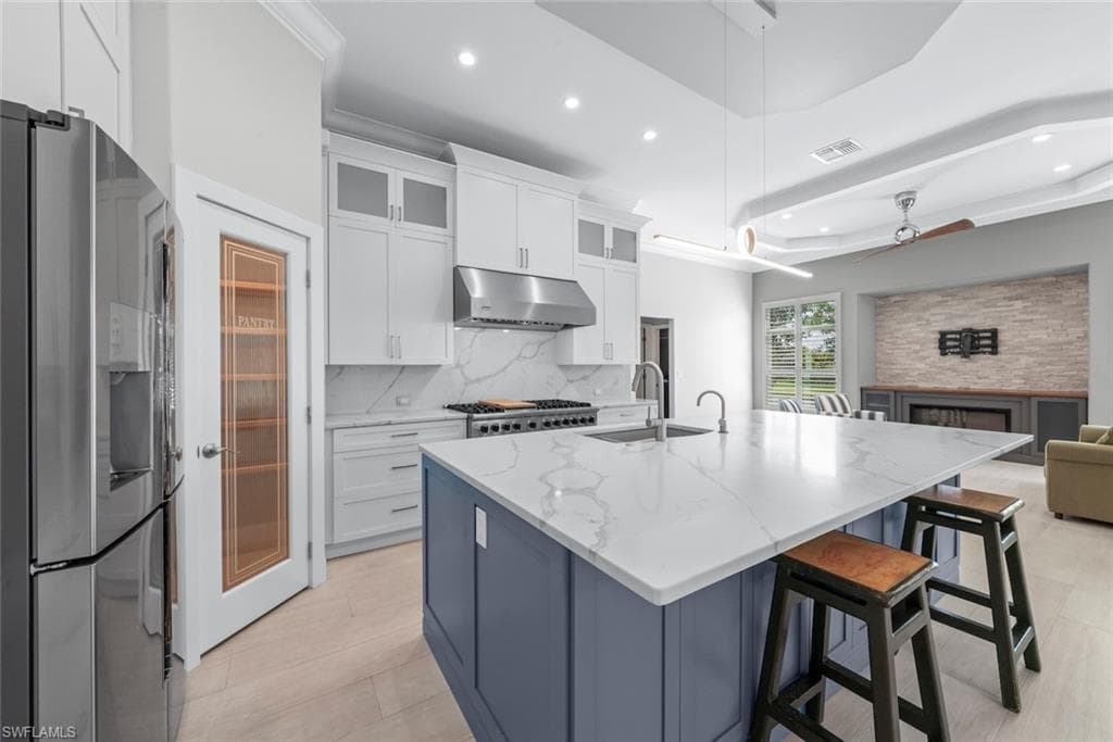 Spacious kitchen featuring a large central island with a quartz countertop, stainless steel appliances, white cabinetry with glass inserts, a dedicated pantry with a glass door, and light wood-finish flooring