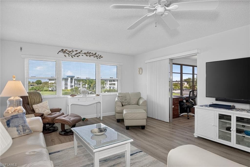 Bright living area featuring wood-finish flooring, a white ceiling fan, and large windows with horizontal blinds