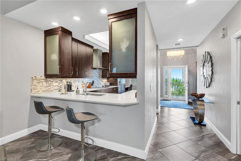 Modern kitchen featuring dark wood cabinetry, frosted glass cabinet fronts, a light-toned tile backsplash, and a breakfast bar with a white countertop