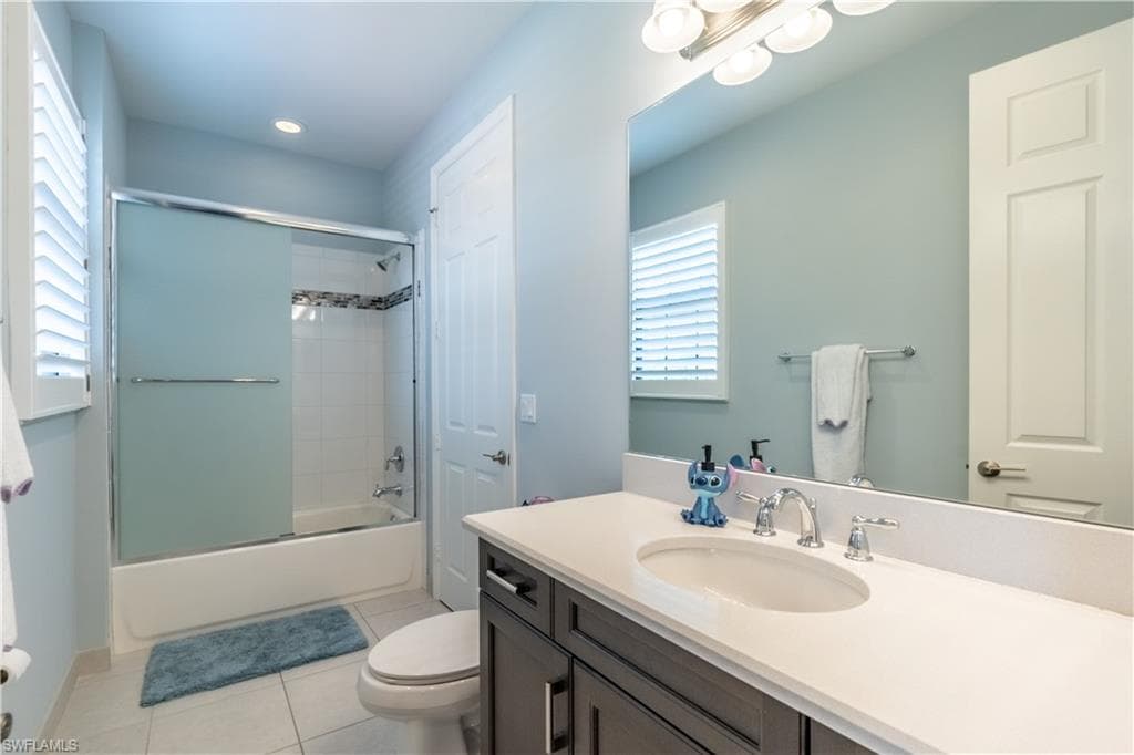 Bathroom featuring a vanity with an undermount sink and light-toned countertop, a shower-tub combination with sliding frosted glass doors, white subway tile surround, and a window with plantation shutters