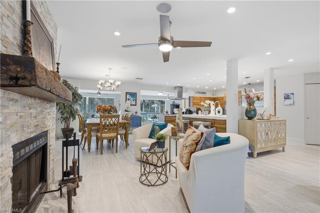 Open-concept living space featuring light wood-finish flooring, a stacked stone fireplace with a thick wood mantel, recessed lighting, and a ceiling fan