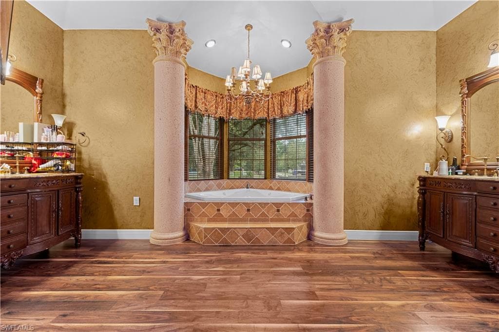 Spacious bathroom featuring a built-in jetted tub with tiled surround, two decorative columns, a chandelier, and wood-finish flooring
