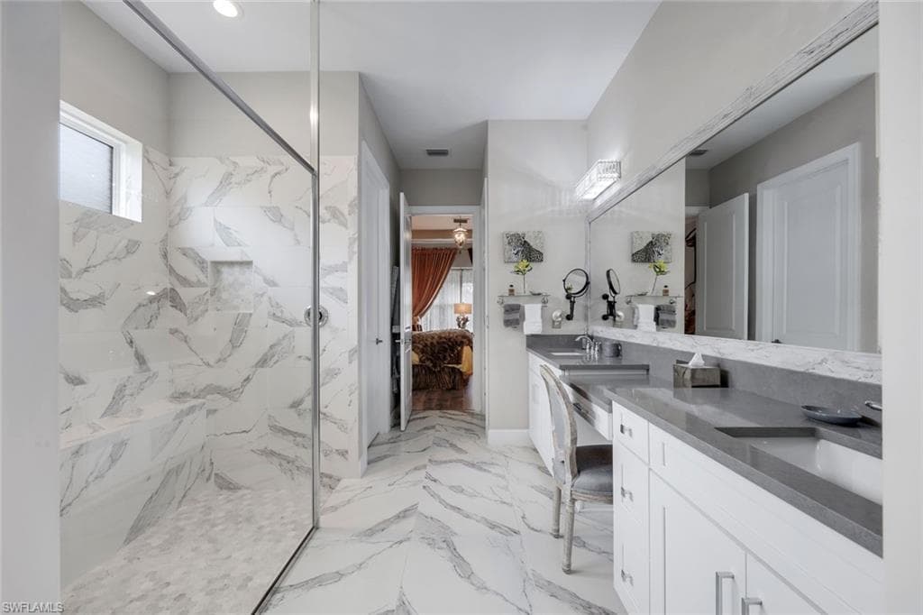 Spacious bath featuring a glass-enclosed shower with a built-in bench, dual vanity with undermount sinks, stone countertops, and full-wall mirror