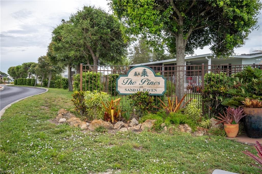 Community entrance sign featuring decorative landscaping, mature trees, and a wrought-iron fence