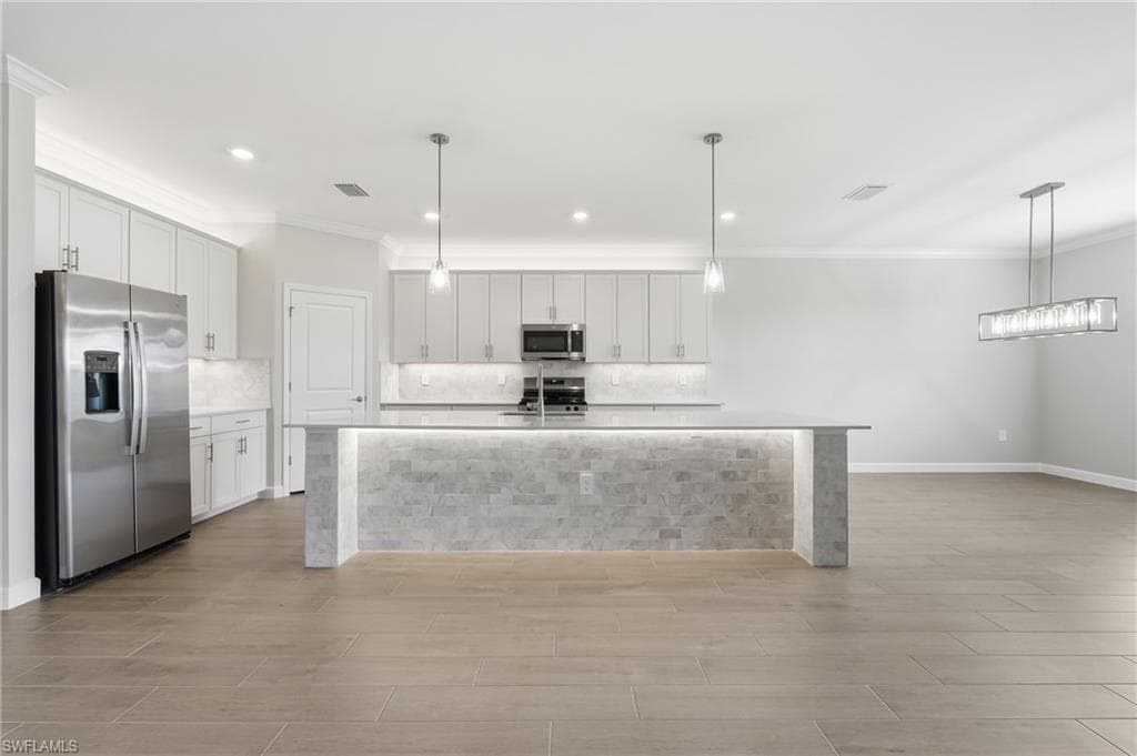 Open-concept kitchen featuring an illuminated island with a stone veneer base, stainless steel appliances, white cabinetry, a light-toned backsplash, and wood-finish flooring