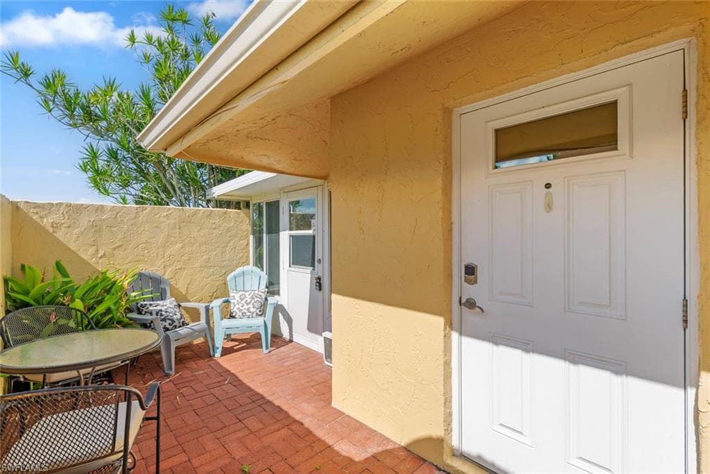 Inviting entry patio featuring a white paneled door with transom window, textured stucco exterior, red brick paving, and a garden bed with lush foliage