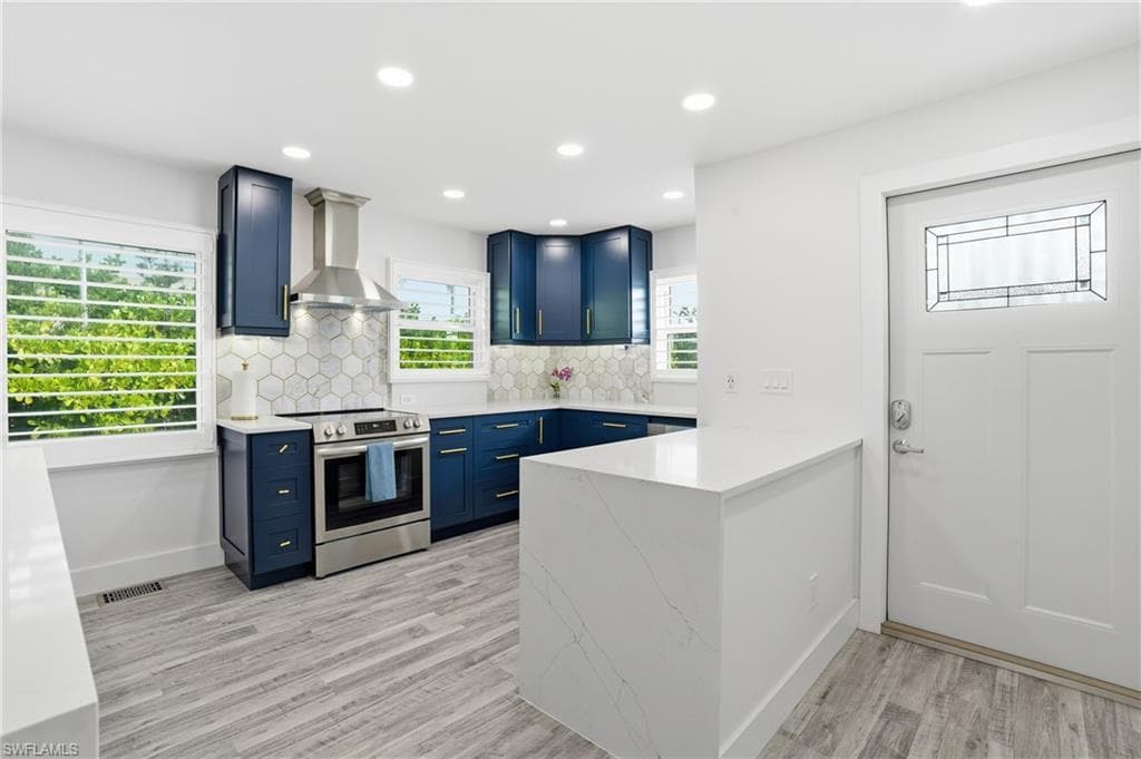 Kitchen with wall chimney exhaust hood, stainless steel electric range, a peninsula, blue cabinets, and light stone countertops - Virtually Edited Image