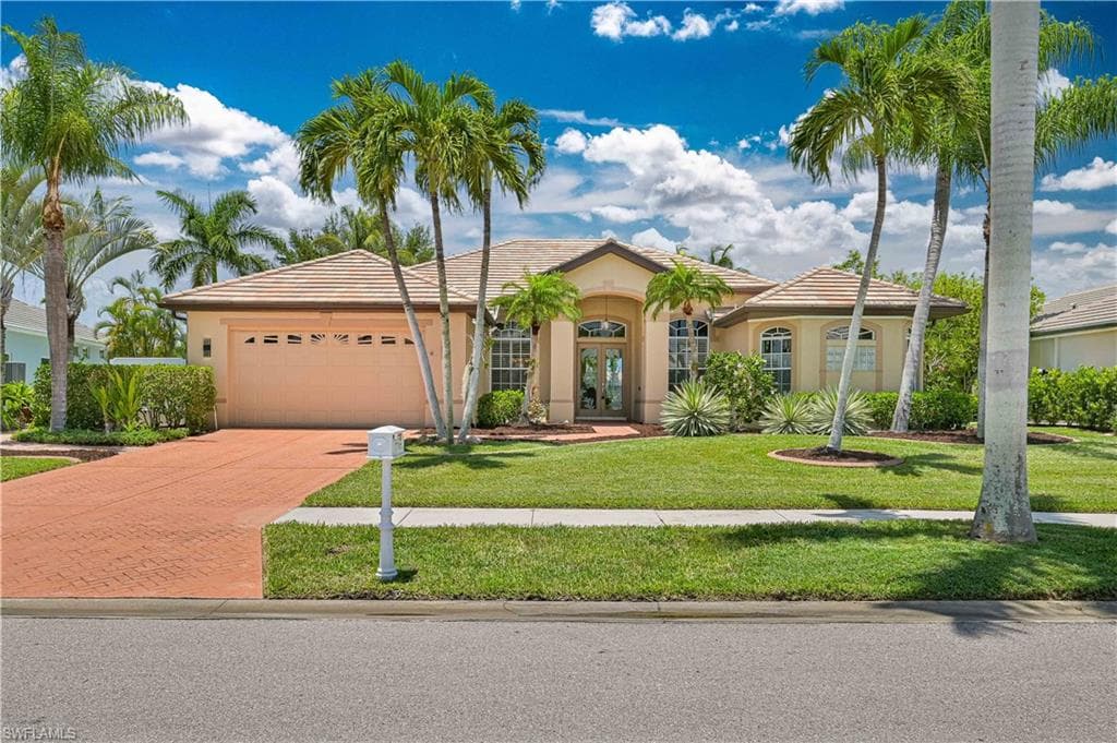 Stucco exterior with a tiled roof, featuring a brick paver driveway, an attached garage, and mature palm trees