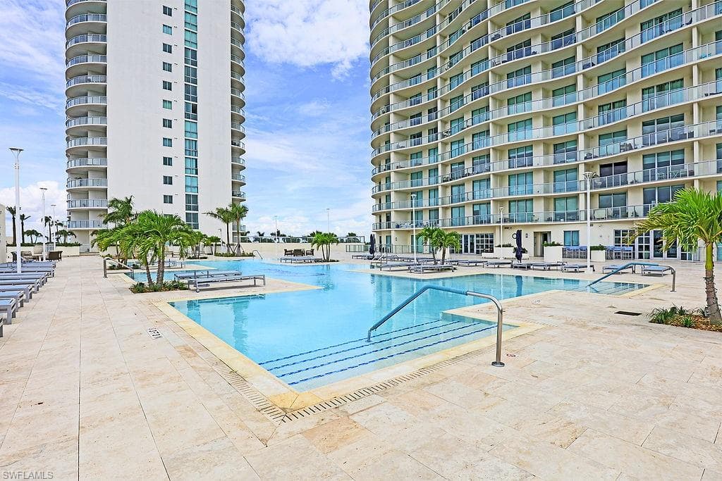 Resort-style pool deck with multiple swimming areas, expansive lounge seating, mature palm trees, and light-colored stone paving, flanked by two high-rise buildings featuring extensive private balconies