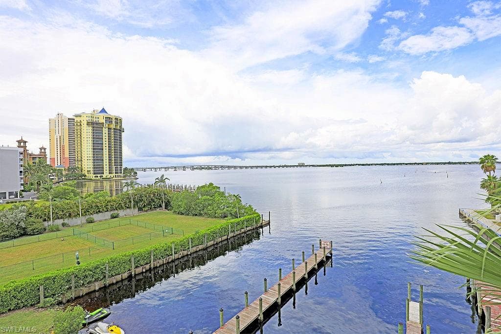 Expansive waterfront setting featuring a multi-story residential building, a long pier extending into the water, and a broad body of water with a distant bridge